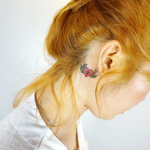 Tatuagem colorida atrás da orelha, um lindo coração com asas, trazendo feminilidade e delicadeza.