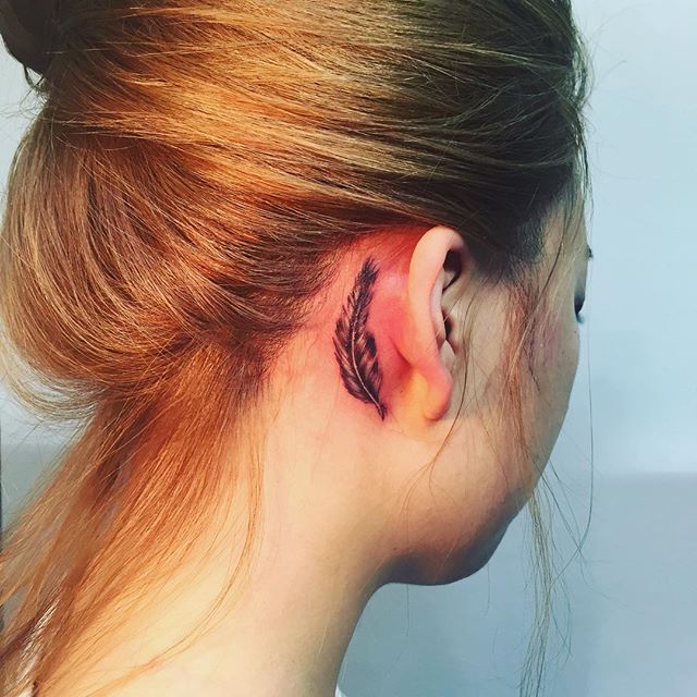 Tatuagem feminina atrás da orelha de uma pena de pássaro.