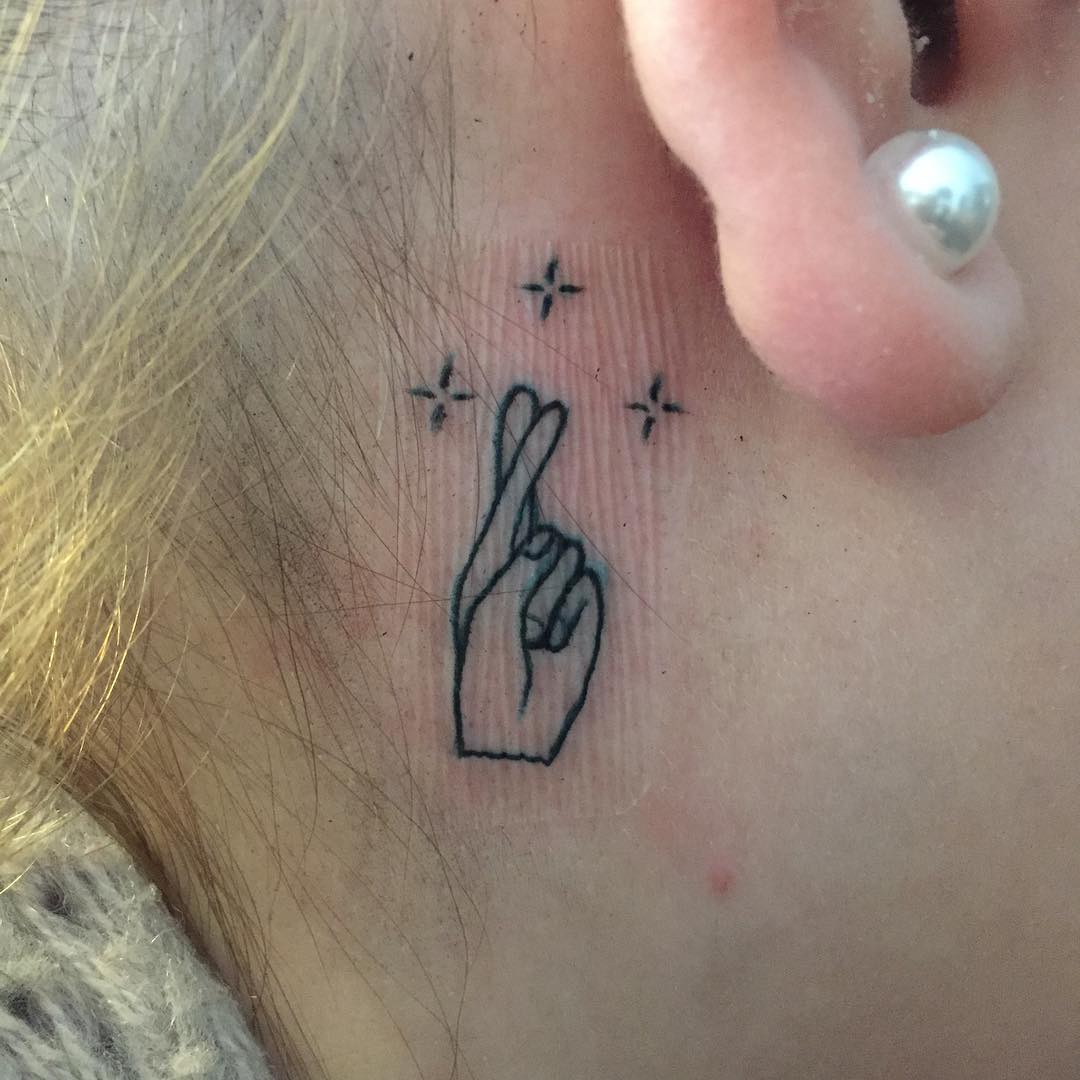 tatuagem de dedos cruzados e estrelas atrás da orelha, para atrair a boa sorte.