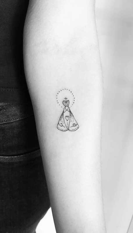 Tatuagem de santa pequena e delicada no braço feminino. A santa de devoção escolhida é Nossa Senhora Aparecida