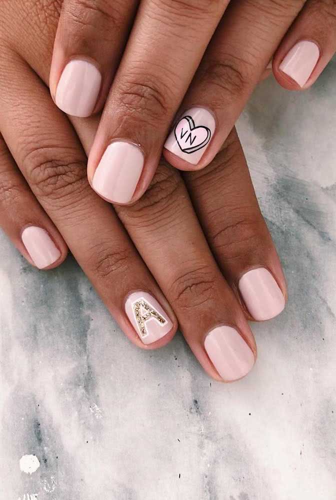 Unhas de noiva curtas pintadas em esmalte rosa clarinho com adesivos de iniciais estilizadas dos noivos na filha única de cada mão.