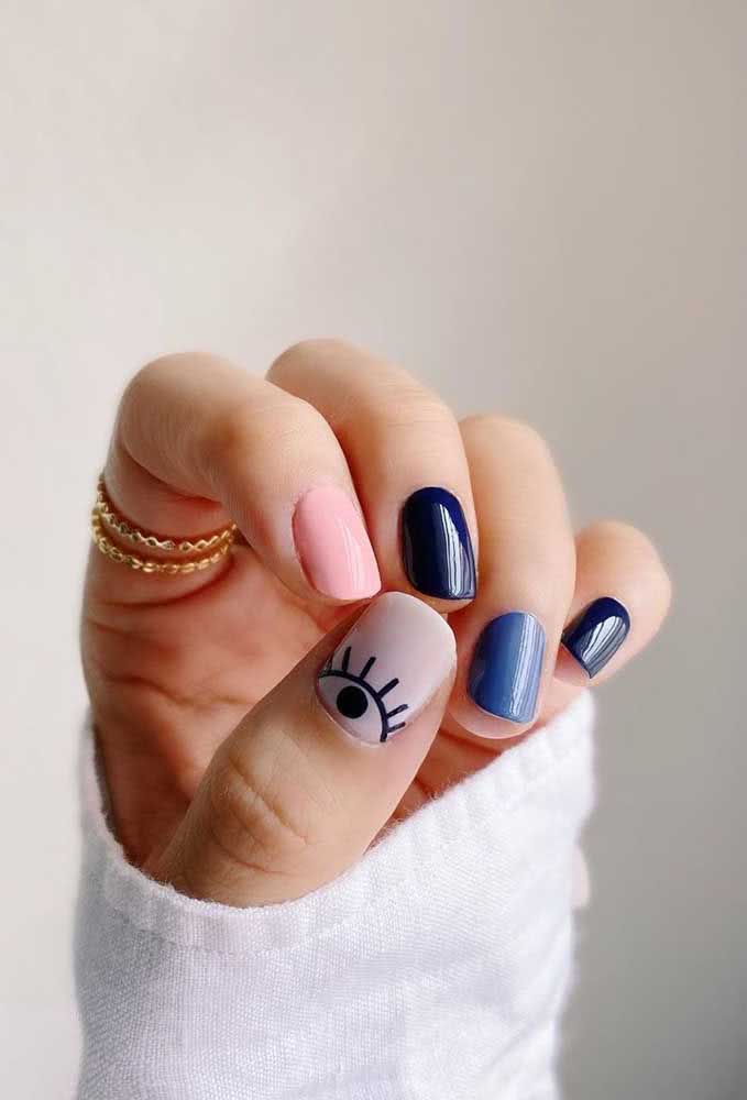 Composição de unhas multicoloridas super jovial e vibrante, com a união de dois tons de azul, rosa clarinho e branco, com desenho de olho estilizado na meia lua do polegar. 