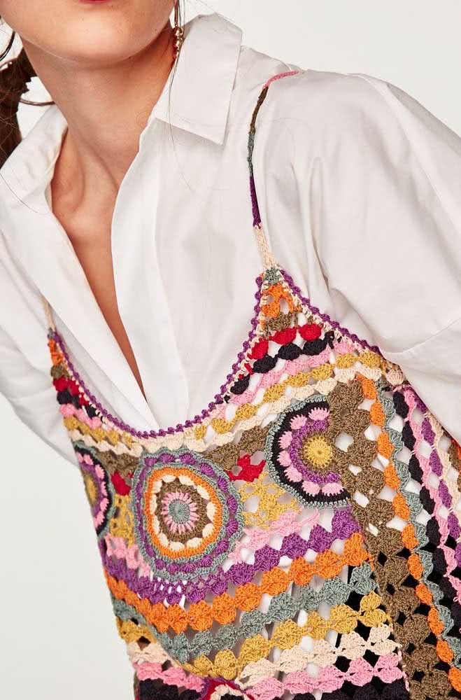 Uma blusa de alcinha de crochê cheia de cor e com padrões desencontrados, usada com camisa branca social ao estilo hi-lo.