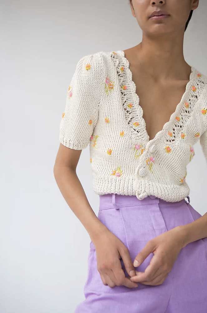 Duas técnicas artesanais em uma peça delicada e aconchegante: um bolero de crochê branco com flores rosas e laranjas bordadas. 
