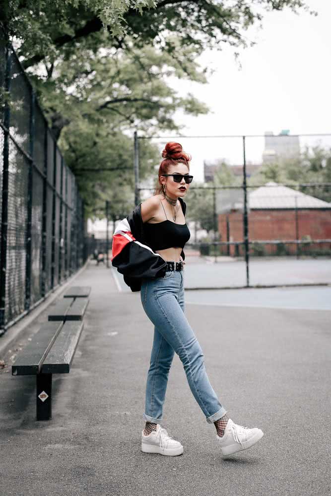 Este look urbano com mom jeans, cinto de rebite, top preto, jaqueta esportiva oversized, meia arrastão e tênis branco é outra ideia que une diversos elementos da moda das décadas passadas num visual punk-rock.