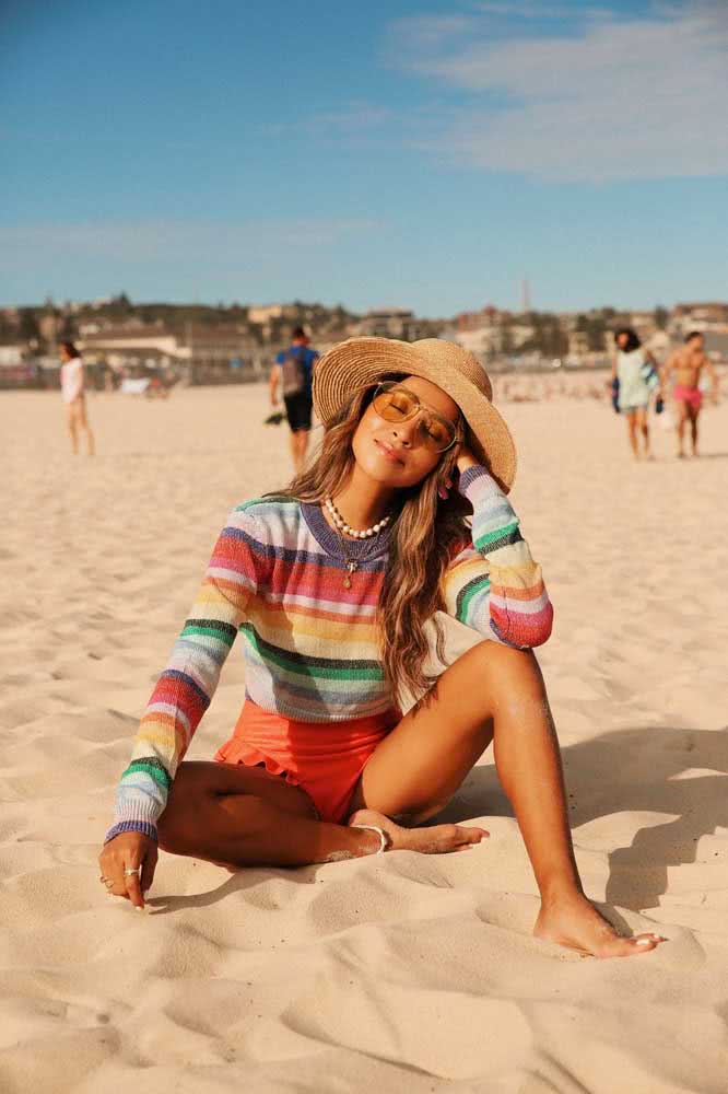 Blusa listrada multicolorida de manga comprida, shorts laranja e chapéu, o look perfeito para quem quer curtir um dia de sol brando na praia.
