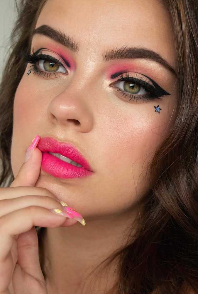 Maquiagem tumblr com delineado gráfico, sombra rosa localizada, cílios realçados, batom rosa e adesivos de estrela para quem busca um visual cheio de atitude.