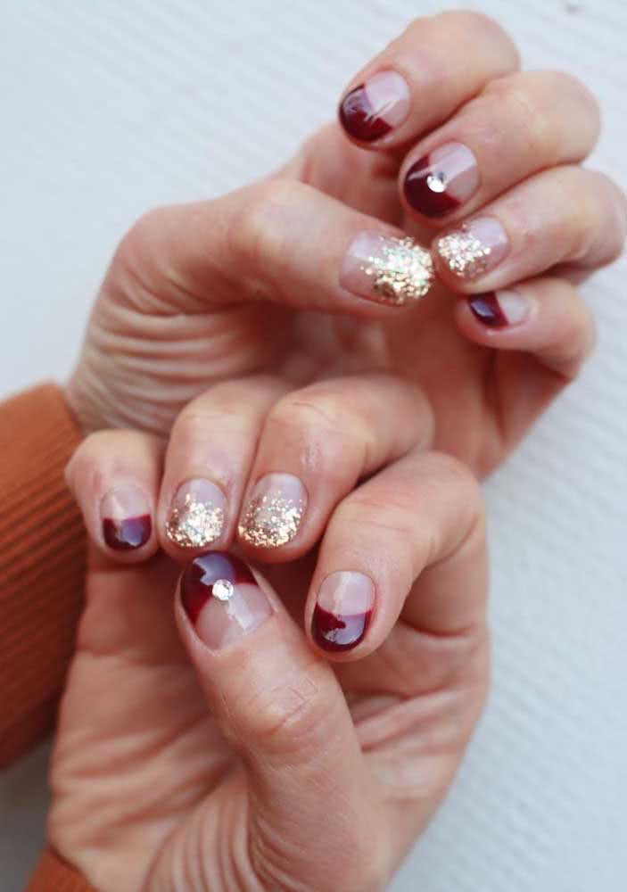 Unhas decoradas com pedras, que trazem mais brilho para a composição em vermelho que contrasta com o glitter dourado. 