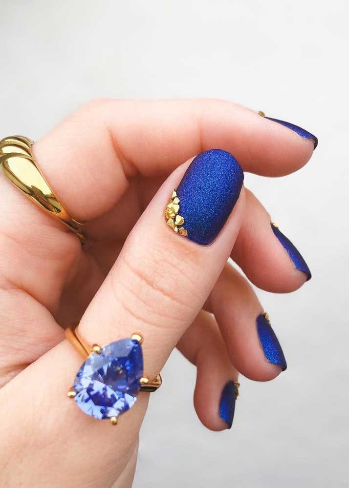 Para combinar com o seu visual, nail art com esmalte fosco com glitter azul anil e um detalhe em pedras douradas.