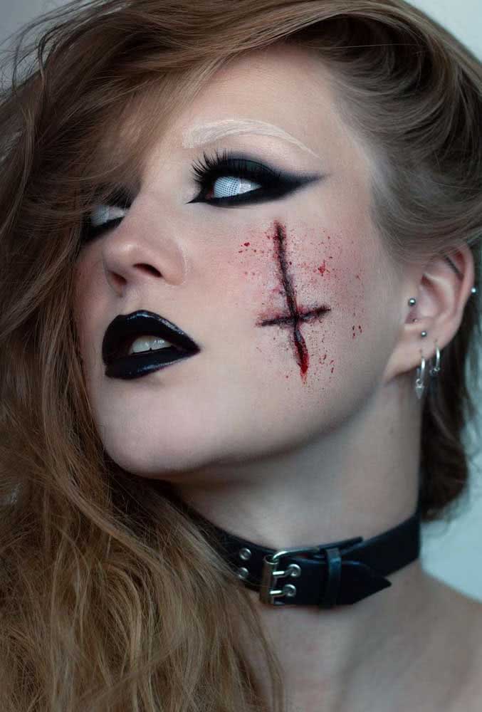 Maquiagem de halloween assustadora de anticristo com direito à cruz invertida com efeito de corte na bochecha, batom e sombra esfumada preta e lentes de contato brancas.