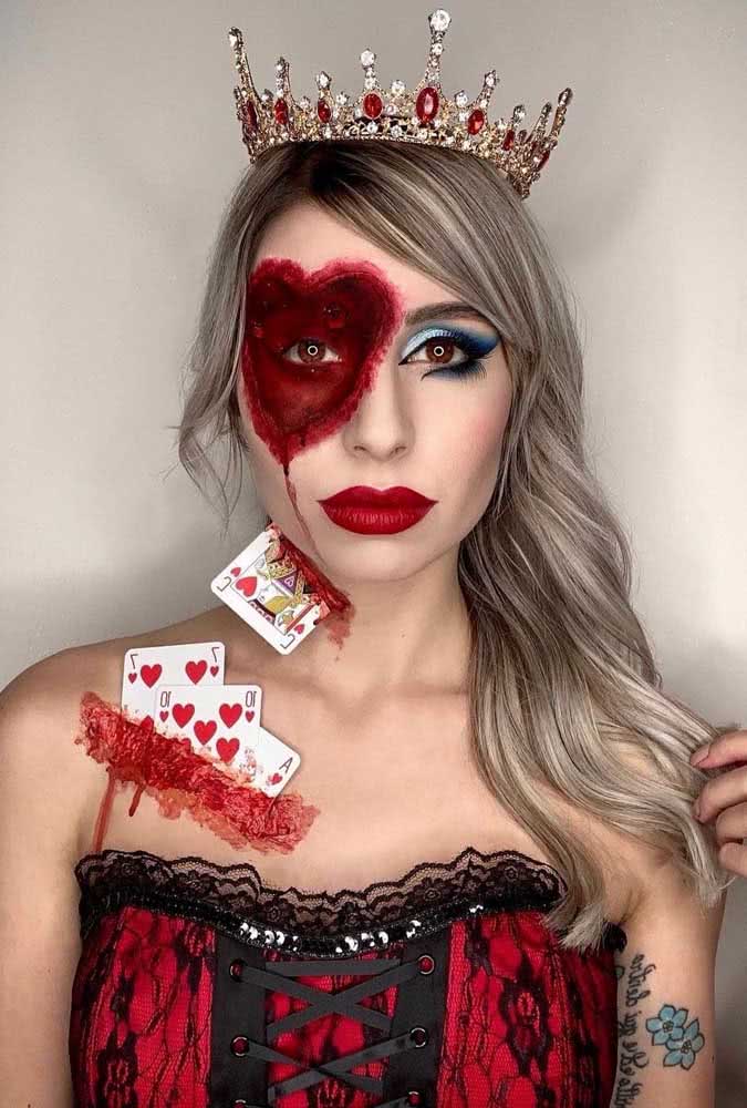 Maquiagem de halloween de rainha de copas macabra com coração ensanguentado ao redor do olho, coroa e cartas de baralho aplicadas.