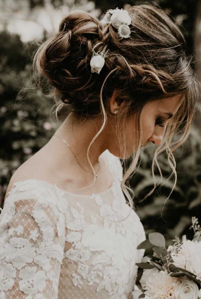 Penteado preso para noiva com coque baixo com topete e trança lateral embutida com algumas mechas soltinhas e flores brancas para um visual romântico. 