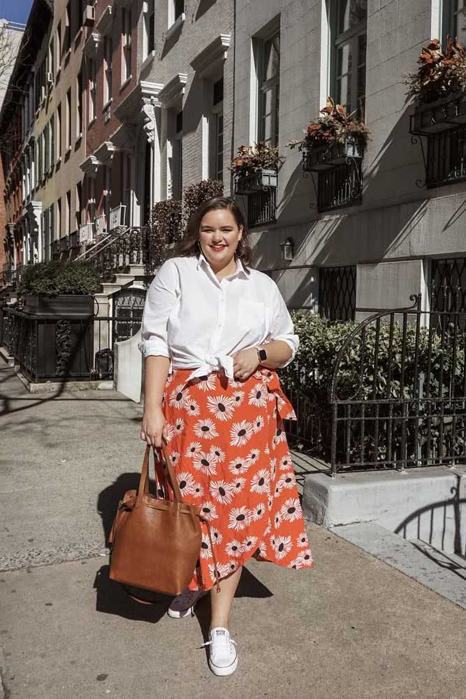 Conforto e muito charme nesta composição com tênis branco, saia midi floral laranja e camisa branca amarrada na cintura. 