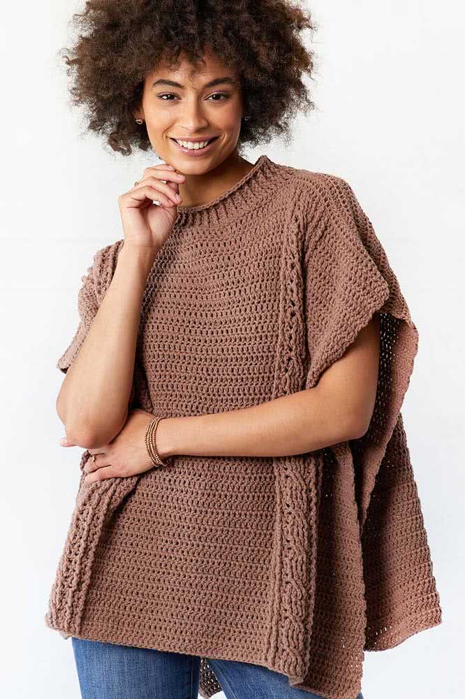 Poncho de crochê marrom com modelo que se assemelha a uma blusa soltinha, com as laterais abertas e um padrão com ponto trançado nas laterais. 