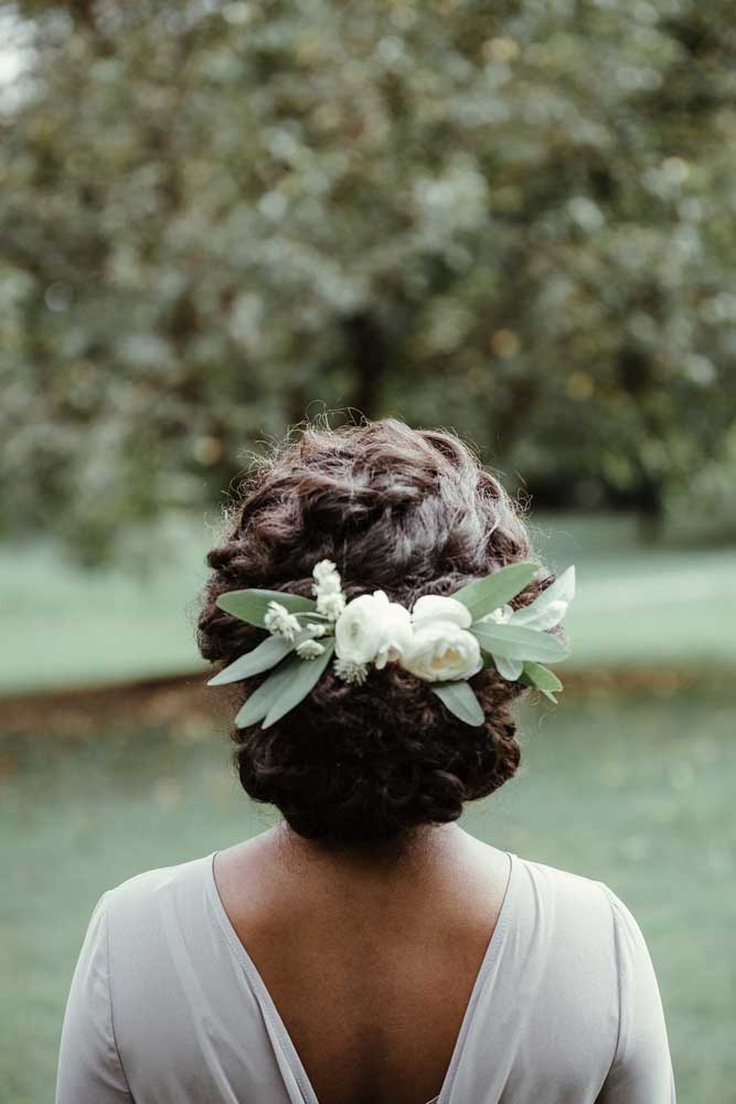 Arranjo de flores brancas e folhas traz um toque a mais de natureza e frescor para este coque em cabelo cacheado para noiva.