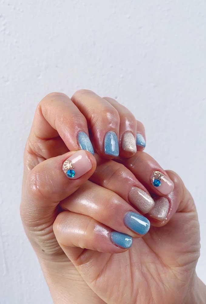 Outra ideia para trazer o mar para a decoração das suas unhas, dessa vez numa composição de esmalte glitter bege e azul e a aplicação de uma pedra e uma concha dourada. 