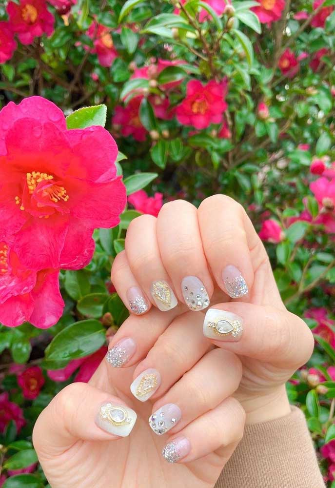 Uma composição cheia de delicadeza e charme com unhas brancas e aplicação de glitter em degradê e de jóias douradas e prateadas.