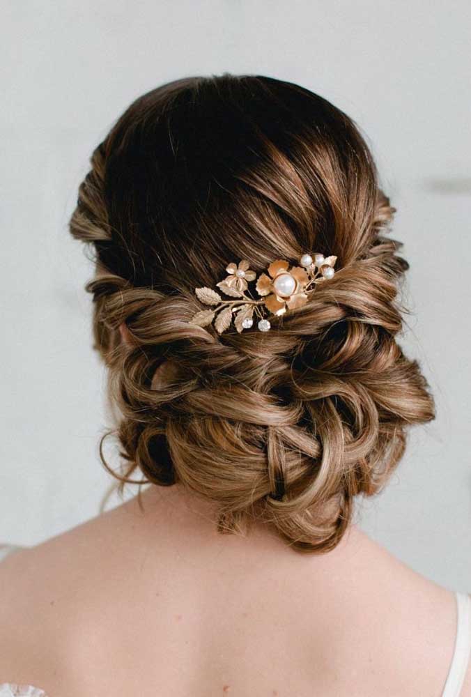 Começando com um penteado para casamento preso em um coque baixo soltinho com as mechas laterais do cabelo torcidas e uma presilha floral decorando.