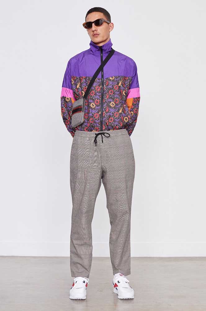 Muito estilo nesse look com roupa anos 80 masculina composta por calça social, sneakers, jaqueta com bastante cor e estampa e uma bolsa transpassada.