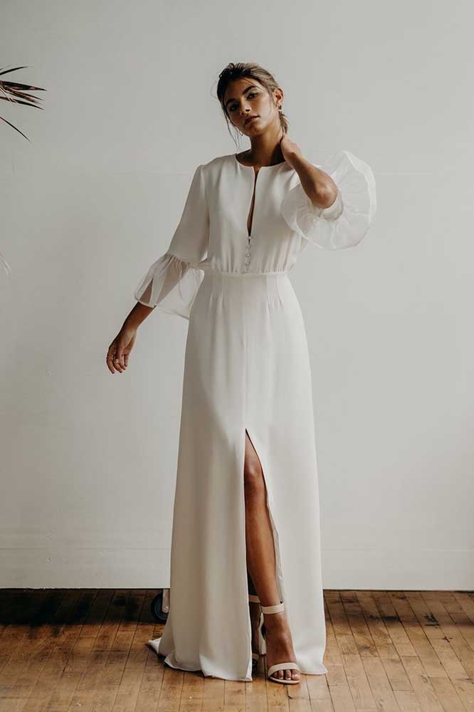Feito num tecido branco liso, esse vestido de noiva evasê longo de manga média ganha uma fenda frontal que garante maiores movimentos e combina com o decote discreto. 