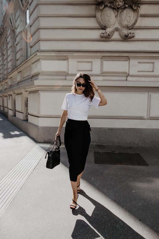 Começando com este look com saia preta envelope em tamanho midi, combinando com uma camiseta branca e sandália, perfeito para o trabalho em dias mais quentes. 