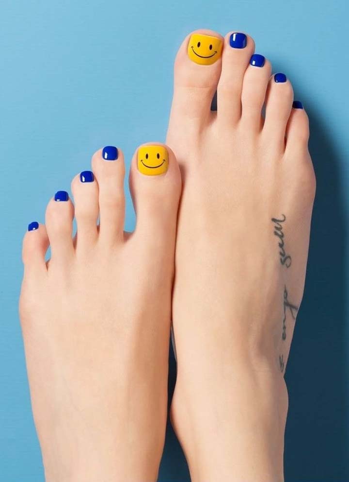 Unhas do pé decoradas em azul anil, exceto a do dedão, que ganha um tom de amarelo canário e uma smiley face bem divertida. 