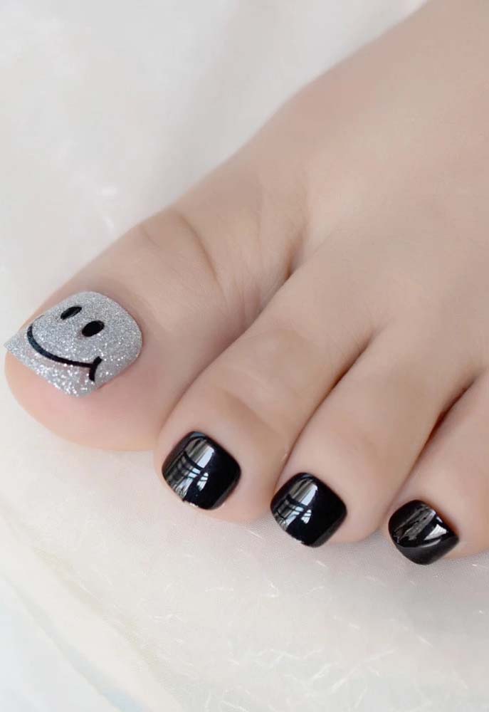Alegre, mas sem perder o estilo de atitude: uma composição de unhas do pé decorada em preto, exceto no dedão, que ganha uma cobertura em glitter cinza e uma smiley face.