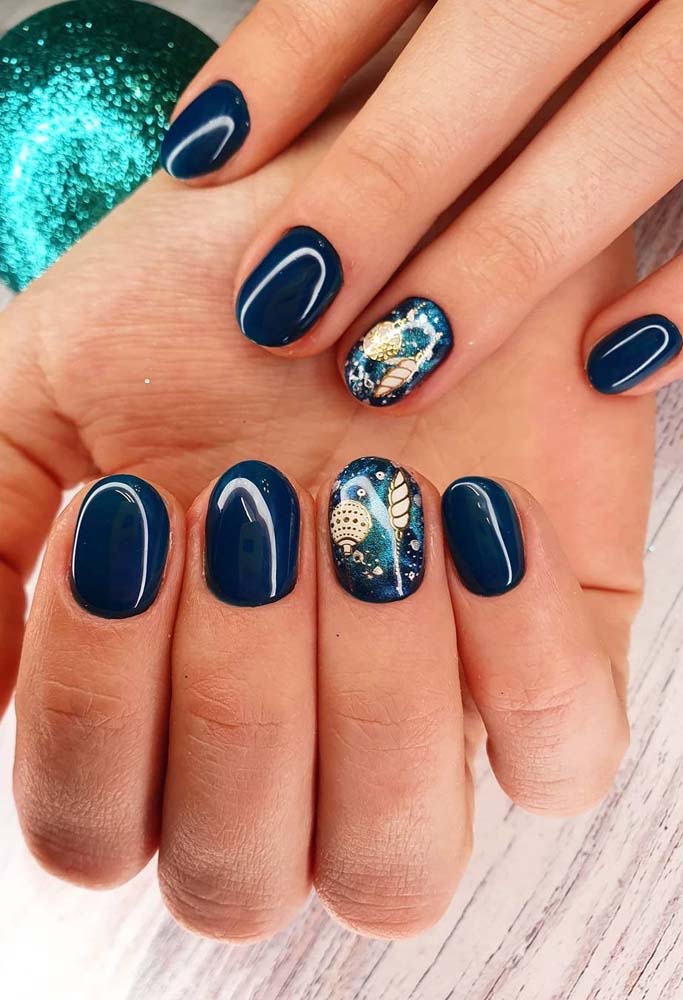 Unhas decoradas azul escuro curtinhas com uma decoração com glitter e adesivos festivos na filha única. 