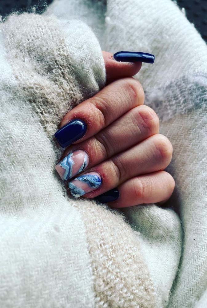 Unhas decoradas azuis escuras com gêmeas em um tom de rosa claro e listras diagonais em azul e branco marmorizados. 