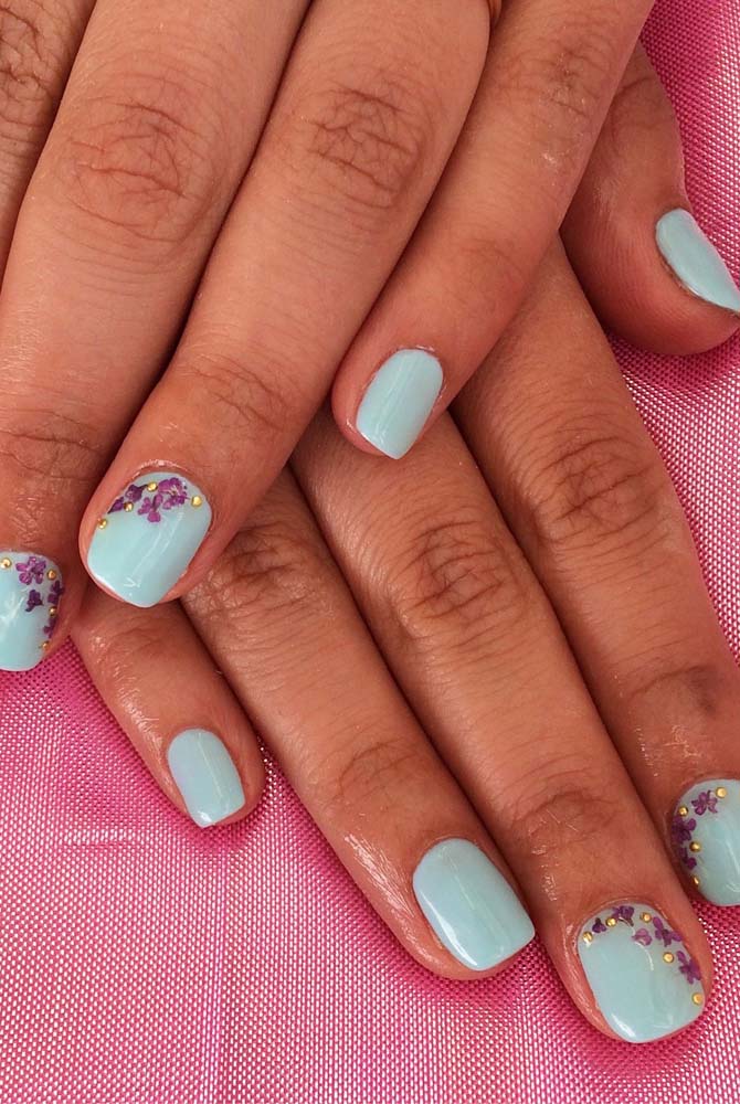 Muita delicadeza nessas unhas decoradas azuis bebê com aplicação de adesivos florais roxos e strass dourados. 