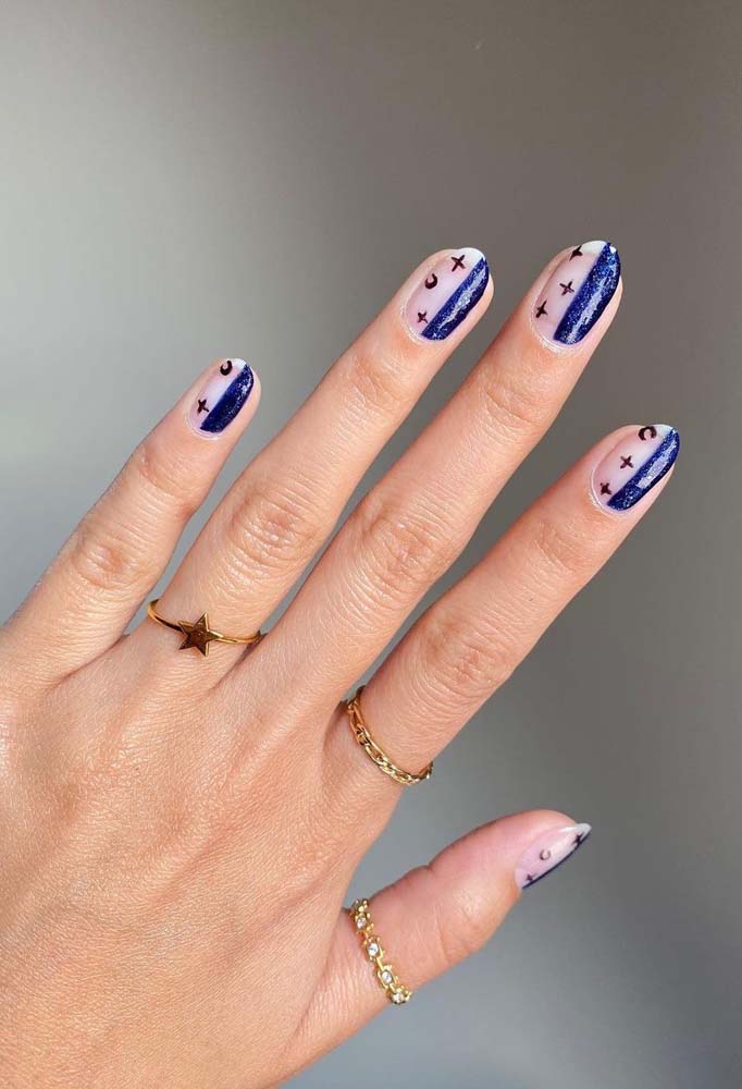 Half nails com tema céu estrelado: de um lado, uma pincelada de azul índigo com toques de branco e de glitter e, do outro, estrelas e luas desenhadas com esmalte preto sobre o tom natural da unha. 
